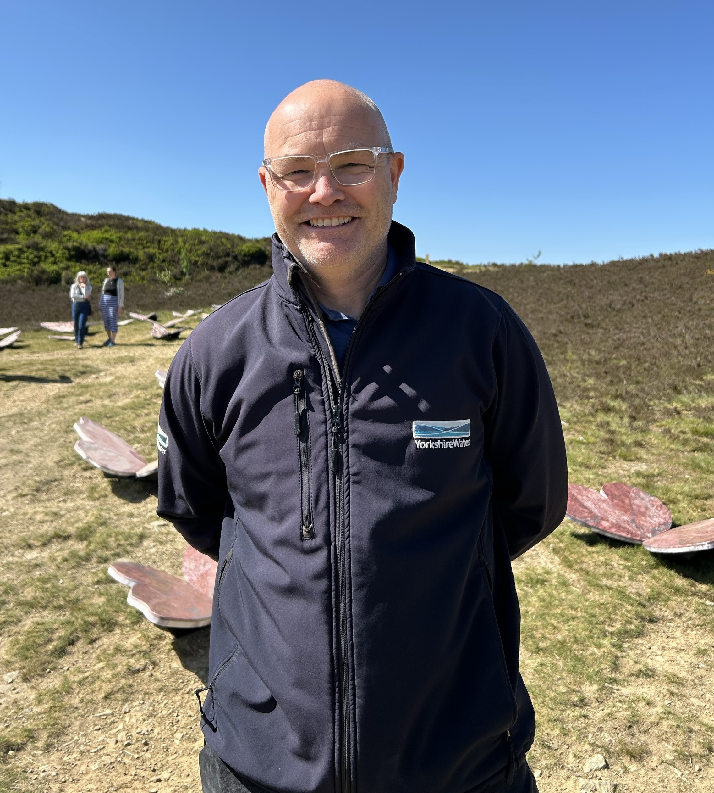 man in blue jacket stands on a hillside with blue sky behind him