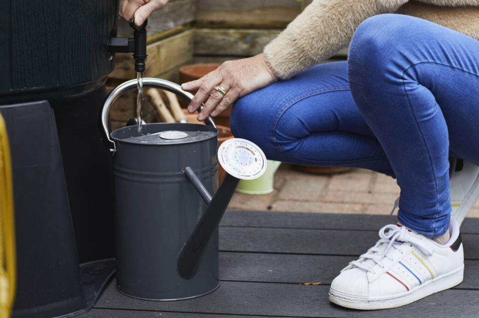 person in jeans kneeling down to fill watering can from a water butt tap